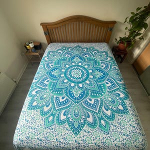 Puede incluir: Una cama con cabecero de madera está cubierta con una colcha con estampado de mandala blanco, turquesa y azul. La colcha presenta un gran diseño floral en el centro, con detalles intrincados y un borde de patrones florales más pequeños.