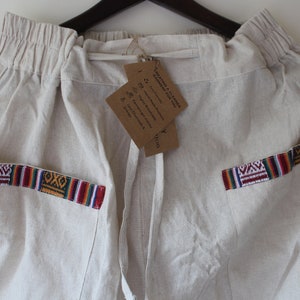 Pode incluir: Calças de linho branco sujo com cós elástico e cordão. Os bolsos apresentam acabamentos tecidos coloridos. Uma etiqueta castanha pende do cordão.