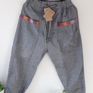 Pode incluir: Calças cinzentas com detalhes estampados coloridos nos bolsos. As calças têm cintura com cordão e punhos elásticos. O tecido parece ser um material tecido. As calças estão penduradas num cabide de madeira.