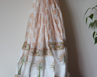 Könnte beinhalten: Weißer Schal mit einem sich wiederholenden orangefarbenen Blumenmuster und einem grünen und orangefarbenen Palmenrand. Der Schal ist mit weißen Pompons am unteren Rand verziert. Der Schal hängt an einem Holzbügel.