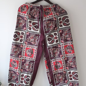 Pode incluir: Calças estampadas em bordô e vermelho com um desenho de elefantes e paisley. As calças estão penduradas em um cabide de madeira. As calças têm cintura com cordão e um ajuste solto. O tecido tem um padrão quadrado.