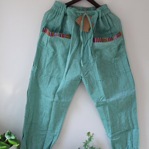 Pode incluir: Calças verde-azuis com cós elástico e cordão. As calças têm dois bolsos com acabamentos tecidos coloridos com padrões vermelhos, laranja e pretos. As calças estão penduradas num cabide de madeira contra uma parede branca.