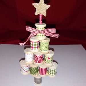 May include: A Christmas tree made of stacked wooden spools wrapped in red, green, and white patterned fabric. The tree is topped with a gold glitter star and a red and white bow.
