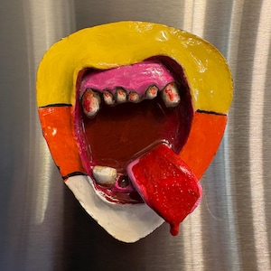 May include: A colorful, abstract sculpture of a mouth with a yellow top, orange sides, and a white bottom. The mouth is open, revealing pink gums, teeth, and a red tongue. The sculpture is mounted on a metallic surface.