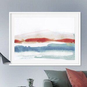 Op de afbeelding: Abstracte aquarelverf in tinten van blauw, groen en rood. Het schilderij toont een landschap met een bergketen op de achtergrond en een waterlichaam op de voorgrond. Het schilderij is ingelijst in een wit frame.