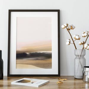 May include: A black framed print of a watercolor painting depicting a beach scene with a soft pink and orange sunset. The print is displayed on a white wall with a vase of cotton flowers and a black bottle on a wooden table.