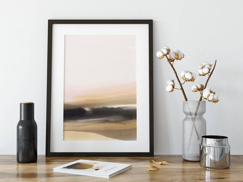 May include: A black framed print of a watercolor painting depicting a beach scene with a soft pink and orange sunset. The print is displayed on a white wall with a vase of cotton flowers and a black bottle on a wooden table.