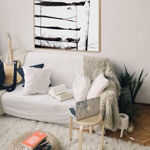 May include: An abstract black and white painting in a wooden frame hangs above a white sofa. The artwork displays horizontal and vertical black brushstrokes. A guitar, books, and a laptop are also visible, creating a comfortable living area.