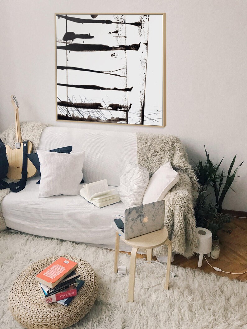 May include: An abstract black and white painting in a wooden frame hangs above a white sofa. The artwork displays horizontal and vertical black brushstrokes. A guitar, books, and a laptop are also visible, creating a comfortable living area.