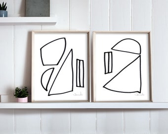 May include: Two black and white abstract geometric line art prints in simple wooden frames. The prints are hung on a white wall with a white shelf underneath.