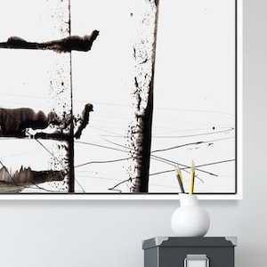May include: Abstract black ink painting on white canvas. The artwork features bold, vertical strokes and splatters, creating a dynamic composition. The painting is framed in a white border. A white vase with pencils sits on a grey box.