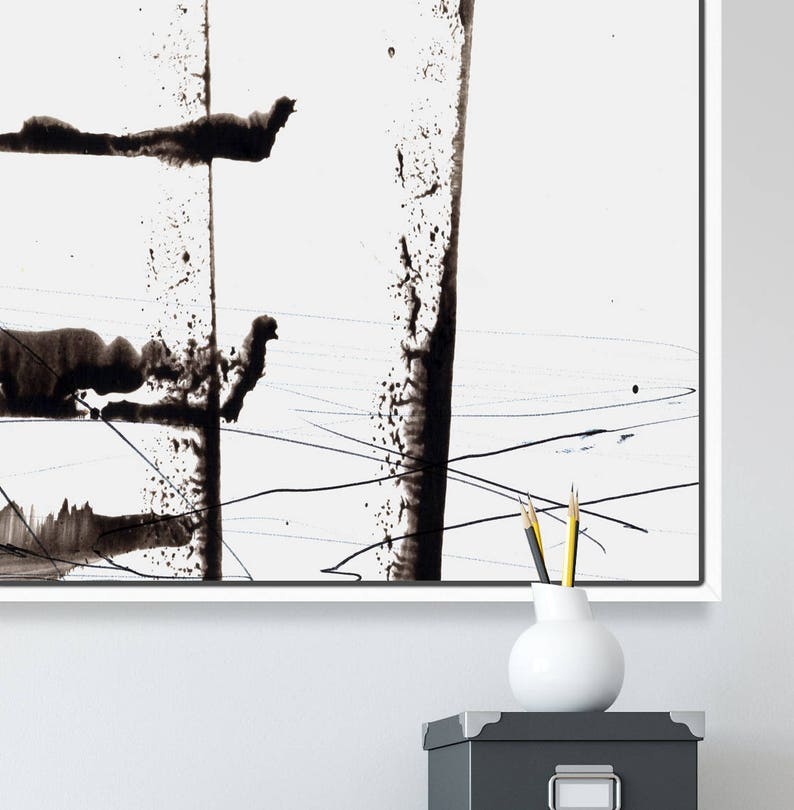 May include: Abstract black ink painting on white canvas. The artwork features bold, vertical strokes and splatters, creating a dynamic composition. The painting is framed in a white border. A white vase with pencils sits on a grey box.