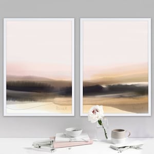 May include: Two framed abstract landscape paintings with a soft pink and brown color scheme. The paintings are in white frames and are hung on a white wall. A white flower in a glass vase and a white mug are on a white table in front of the paintings.