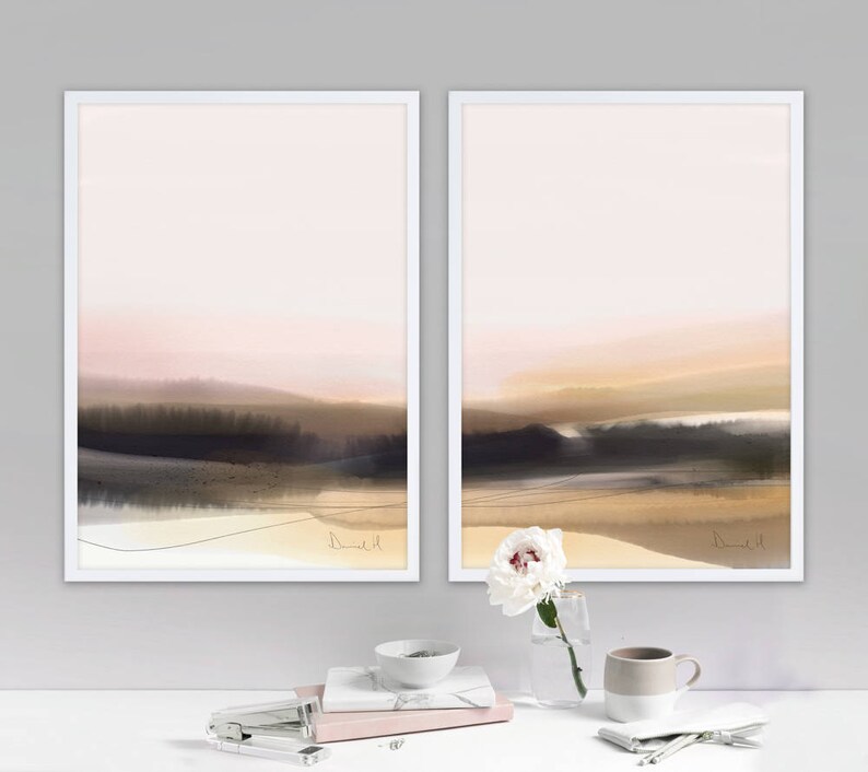 May include: Two framed abstract landscape paintings with a soft pink and brown color scheme. The paintings are in white frames and are hung on a white wall. A white flower in a glass vase and a white mug are on a white table in front of the paintings.