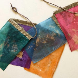 May include: A colorful banner composed of rectangular fabric flags in shades of teal, purple, blue, orange, and pink. Each flag is trimmed with gold-colored material and strung together with a brown cord. The flags have a textured, translucent appearance.