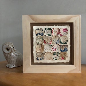 May include: A framed artwork featuring a collage of vintage paper labels in various colors and patterns, arranged in a grid pattern. The labels are slightly faded and have a distressed look, giving the piece a vintage aesthetic.