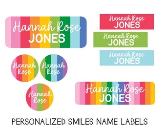 May include: Rainbow-striped name labels with the text "Hannah Rose JONES" in white. There are five labels in total, three are rectangular and two are round. The labels are designed for personalizing items.