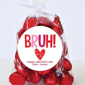 May include: A white circle sticker with red and pink text that says "BRUH!" with a red heart and the text "happy valentine's day from - hunter". The sticker is on a clear plastic bag filled with red foil-wrapped chocolate kisses.