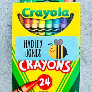 Puede incluir: Una caja de 24 crayones Crayola con una etiqueta personalizada que dice "HADLEY JONES CRAYONS". La etiqueta presenta una abeja de dibujos animados con una sonrisa. La caja es amarilla con detalles verdes y la palabra "Crayola" en letras rojas.