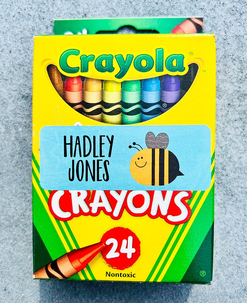 Puede incluir: Una caja de 24 crayones Crayola con una etiqueta personalizada que dice "HADLEY JONES CRAYONS". La etiqueta presenta una abeja de dibujos animados con una sonrisa. La caja es amarilla con detalles verdes y la palabra "Crayola" en letras rojas.