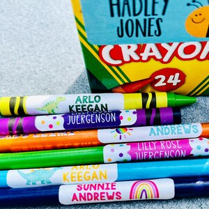 Puede incluir: Una caja de 24 crayones con una etiqueta que dice "Crayola" y una abeja con una cara sonriente. Hay varios crayones con etiquetas personalizadas que dicen "Arlo Keegan Juergenson", "Sunnie Andrews" y "Lilly Rose Juergenson".
