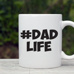 Op de afbeelding: Witte keramische mok met een zwarte hashtag en de tekst "#DAD LIFE" gedrukt op de voorkant.