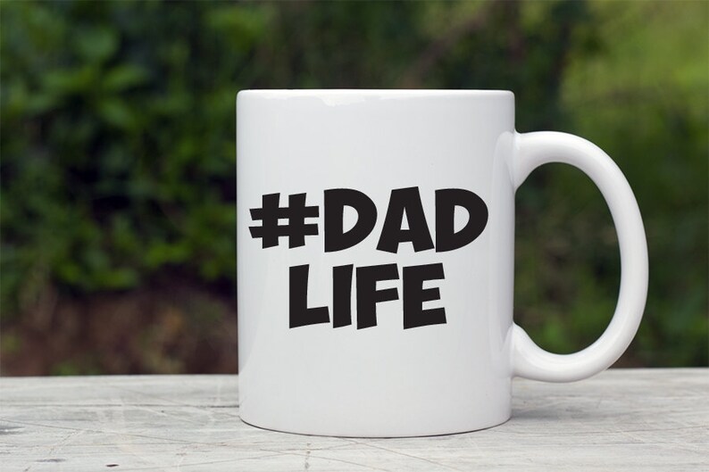 Op de afbeelding: Witte keramische mok met een zwarte hashtag en de tekst "#DAD LIFE" gedrukt op de voorkant.