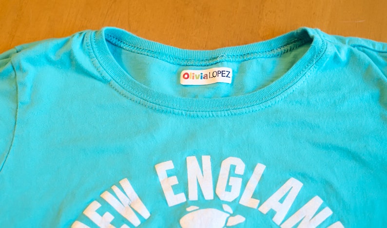 Op de afbeelding: Een turquoise blauw t-shirt met een witte afbeelding waarop "NEW ENGLAND" staat en een witte afbeelding van een kreeft. Het shirt heeft een wit label dat in de hals is genaaid met de tekst "Olivia LOPEZ".