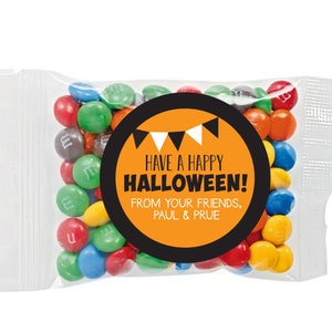 Op de afbeelding: Een kleine doorzichtige plastic zak gevuld met kleurrijke M&M's snoepjes. Een ronde sticker met zwarte en oranje randen en witte tekst luidt "Have a Happy Halloween! From your friends, Paul & Prue."