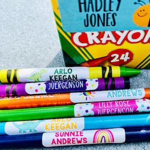 Op de afbeelding: Een doos met 24 kleurpotloden met een label waarop "Crayola" staat en een bij met een smiley. Er zijn verschillende kleurpotloden met gepersonaliseerde labels waarop "Arlo Keegan Juergenson", "Sunnie Andrews" en "Lilly Rose Juergenson" staat.