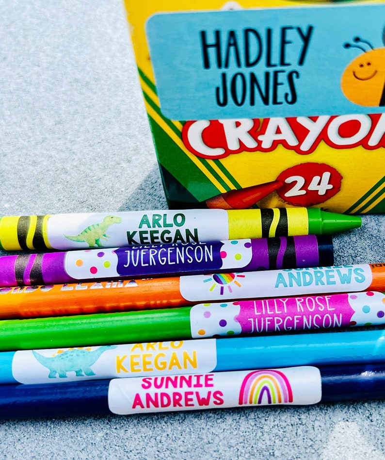 Op de afbeelding: Een doos met 24 kleurpotloden met een label waarop "Crayola" staat en een bij met een smiley. Er zijn verschillende kleurpotloden met gepersonaliseerde labels waarop "Arlo Keegan Juergenson", "Sunnie Andrews" en "Lilly Rose Juergenson" staat.