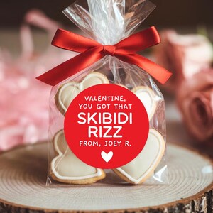 May include: A red circle sticker with white text that reads "Valentine, you got that Skibidi Rizz from, Joey R." with a small white heart in the bottom right corner. The sticker is on a clear plastic bag filled with heart-shaped cookies.