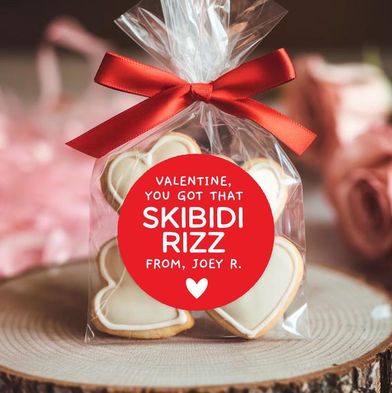 May include: A red circle sticker with white text that reads "Valentine, you got that Skibidi Rizz from, Joey R." with a small white heart in the bottom right corner. The sticker is on a clear plastic bag filled with heart-shaped cookies.