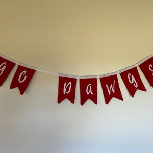 Può includere: Un banner rosso con testo bianco che recita "go dawgs". Il banner è composto da singoli stendardi collegati da un nastro bianco. Gli stendardi sono di colore rosso intenso.