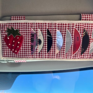 May include: A red and white gingham CD visor organizer with a strawberry applique. The organizer holds several CDs, including one with "PEARL JAM" printed on it. The visor is attached to a car's sun visor.