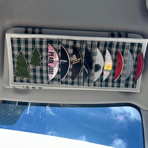 Könnte beinhalten: Ein karierter CD-Visier-Organizer mit weißem Rand, der mehrere CDs enthält. Der Organizer hat zwei grüne Weihnachtsbaum-Applikationen. Eine CD ist mit "PEARL JAM" beschriftet.