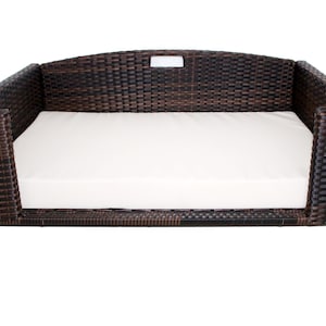 May include: A brown wicker dog bed with a white cushion. The bed has a rectangular shape with raised sides and a curved back. The wicker is woven in a tight pattern, and the cushion appears to be made of a soft fabric.