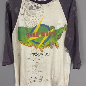 Puede incluir: Una camiseta vintage blanca, verde y amarilla de la banda The Who con un mapa de los Estados Unidos y el texto "Tour '80". La camiseta tiene un diseño de estilo béisbol morado y blanco con agujeros y roturas por todas partes.