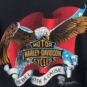 May include: Black t-shirt with a red, white, and blue American flag design. A bald eagle with outstretched wings is flying over the flag. The flag is wrapped around a Harley-Davidson logo with the text "MOTOR HARLEY-DAVIDSON CYCLES". The text "REBEL WITH A CAUSE" is written in a banner below the logo. The text "1987 Harley Davidson, Inc. Mfd by Speed Limit Security" is printed in the bottom right corner.