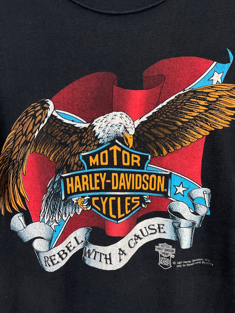 May include: Black t-shirt with a red, white, and blue American flag design. A bald eagle with outstretched wings is flying over the flag. The flag is wrapped around a Harley-Davidson logo with the text "MOTOR HARLEY-DAVIDSON CYCLES". The text "REBEL WITH A CAUSE" is written in a banner below the logo. The text "1987 Harley Davidson, Inc. Mfd by Speed Limit Security" is printed in the bottom right corner.