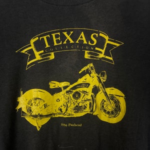Puede incluir: Camiseta negra con un gr&aacute;fico amarillo de una motocicleta Panhead de 1956. El gr&aacute;fico incluye el texto "Texas Collection".