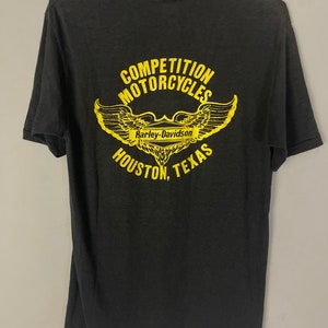 Puede incluir: Camiseta negra con un gr&aacute;fico amarillo de una motocicleta Harley-Davidson con alas y el texto "Competition Motorcycles Houston, Texas".