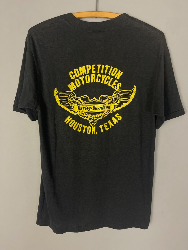 Puede incluir: Camiseta negra con un gr&aacute;fico amarillo de una motocicleta Harley-Davidson con alas y el texto "Competition Motorcycles Houston, Texas".