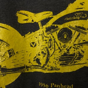 Puede incluir: Una camiseta negra con una imagen dorada de una motocicleta Harley-Davidson Panhead de 1956. La motocicleta est&aacute; delineada en oro y tiene el texto "Harley-Davidson" en el lateral.