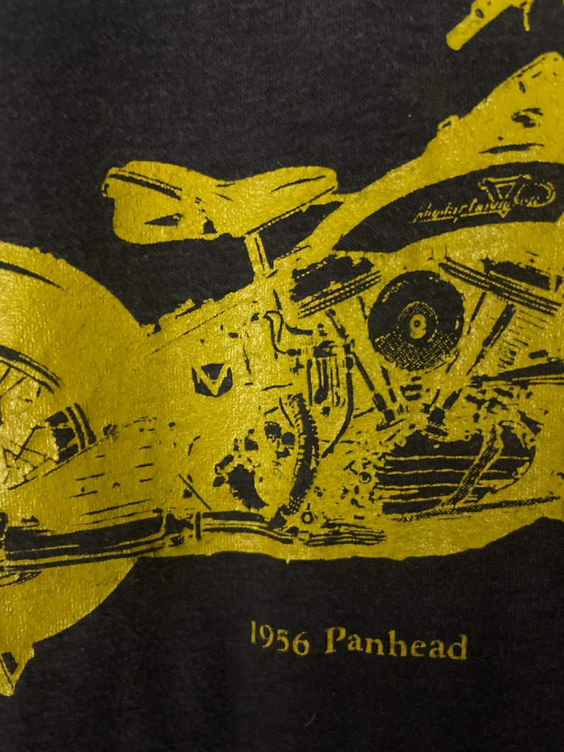 Puede incluir: Una camiseta negra con una imagen dorada de una motocicleta Harley-Davidson Panhead de 1956. La motocicleta est&aacute; delineada en oro y tiene el texto "Harley-Davidson" en el lateral.