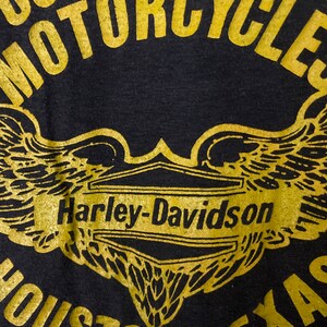 Puede incluir: Camiseta negra con un logotipo dorado de Harley-Davidson y alas. El texto "Competition Motorcycles" est&aacute; por encima del logotipo y "Houston, Texas" est&aacute; por debajo del logotipo.