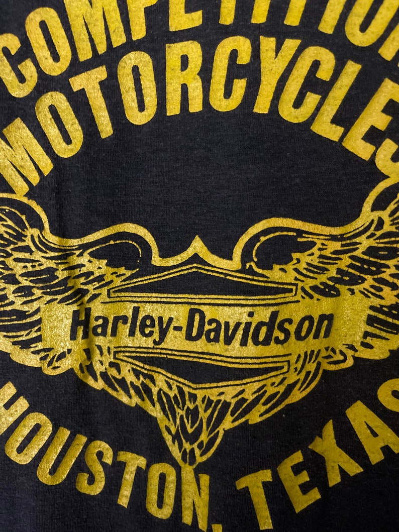 Puede incluir: Camiseta negra con un logotipo dorado de Harley-Davidson y alas. El texto "Competition Motorcycles" est&aacute; por encima del logotipo y "Houston, Texas" est&aacute; por debajo del logotipo.