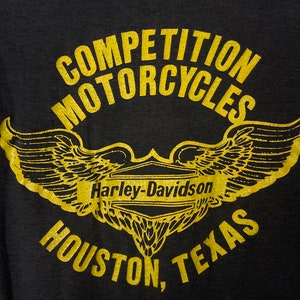 Puede incluir: Camiseta negra con un gr&aacute;fico dorado de una motocicleta Harley-Davidson con alas. El texto "Competition Motorcycles" est&aacute; por encima de la motocicleta y "Houston, Texas" est&aacute; por debajo.
