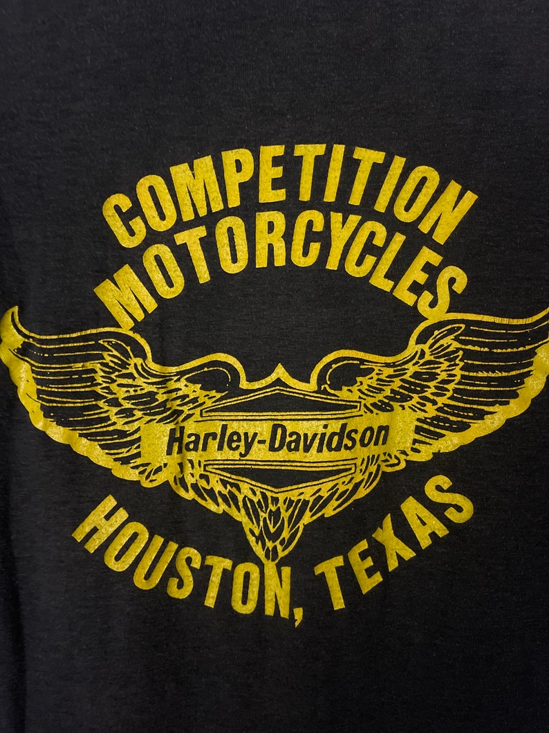 Puede incluir: Camiseta negra con un gr&aacute;fico dorado de una motocicleta Harley-Davidson con alas. El texto "Competition Motorcycles" est&aacute; por encima de la motocicleta y "Houston, Texas" est&aacute; por debajo.