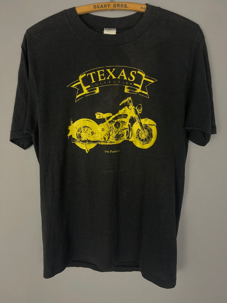 Puede incluir: Camiseta negra con un gr&aacute;fico amarillo de una motocicleta y el texto "Texas Collection" con una pancarta sobre la motocicleta.